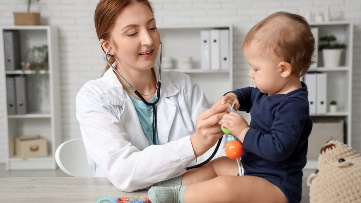 young happy female pediatrician with stethoscope examining cute little baby on table in clinic