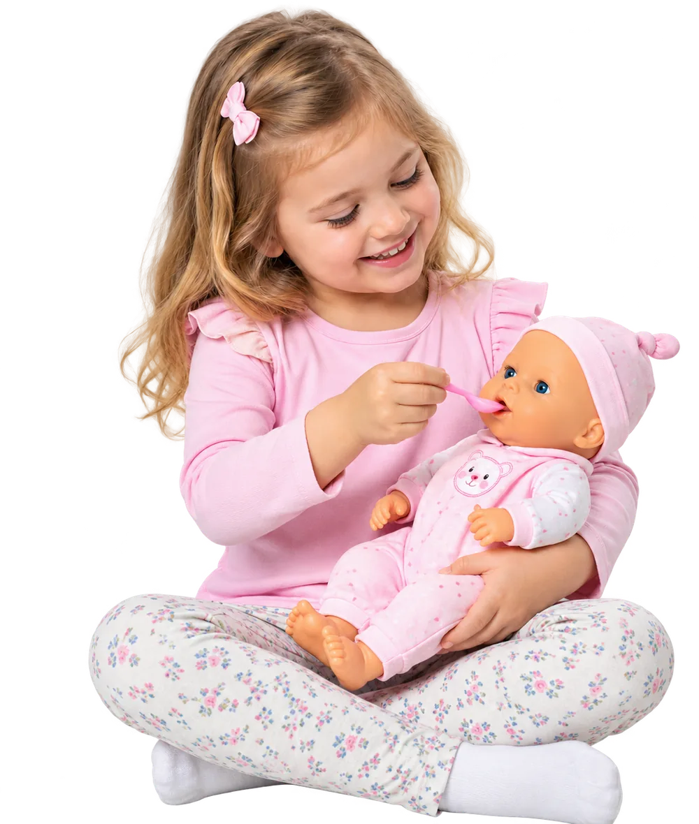 A smiling young girl sitting cross-legged, pretend-feeding her baby doll with a toy spoon